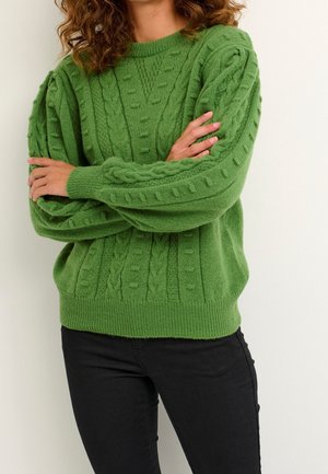 Personne aux bras croisés portant un pull vert en maille torsadée et un pantalon noir devant un fond clair uni.