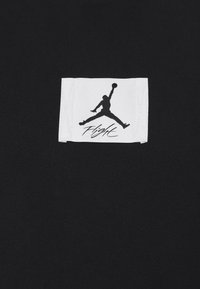 Černá tkanina s tkanou texturou má bílou štítek s černým logem Jumpman a slovem "Flight" v kurzívním písmu.