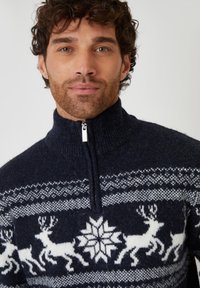 Marineblauer Wollpullover mit hohem Kragen und halbem Reißverschluss. Mit weißen Rentier- und Schneeflockenmuster über der Brust und den Schultern.