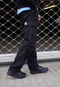 Černé cargo kalhoty mají volný střih, pevnou bavlněnou látku, více kapes a nášivku s logem Carhartt. V kombinaci s černými teniskami.