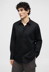 Νεαρός άνδρας με σγουρά μαλλιά, φορώντας μαύρο πουκάμισο με κουμπιά και γκρι παντελόνι με πλαϊνή τσέπη, κοιτάζει προς τα αριστερά σε φόντο απλής επιφάνειας.