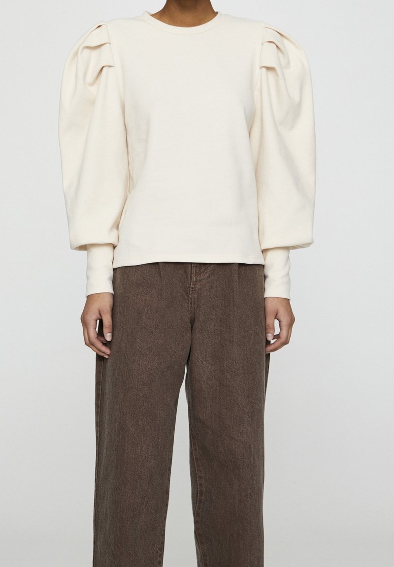 Personne portant un haut crème à manches longues avec des épaules bouffantes et un pantalon marron ample sur un fond clair uni.