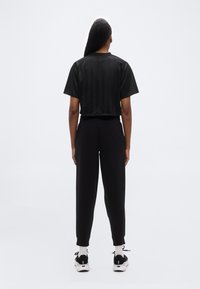 Άτομο που στέκεται, με την πλάτη στην κάμερα, φοράει μαύρο κοντομάνικο μπλουζάκι, μαύρο παντελόνι, άσπρες κάλτσες και μαύρα-άσπρα αθλητικά παπούτσια.