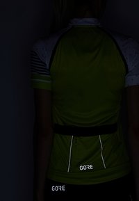 Jersey de ciclismo de manga corta en verde neón con acentos reflectantes, con una cinta negra en la cintura y logotipos blancos, colocado sobre un fondo oscuro.