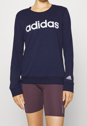Osoba ubrana w granatową bluzę Adidas z białym logo i bordowe szorty Adidas Techfit, stojąca na tle jasnego, jednolitego tła.