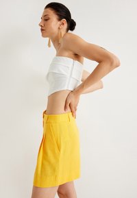 Witte crop top met gelaagd ontwerp en rits aan de achterkant, gecombineerd met een gele linnen mini rok met plooien aan de voorkant.