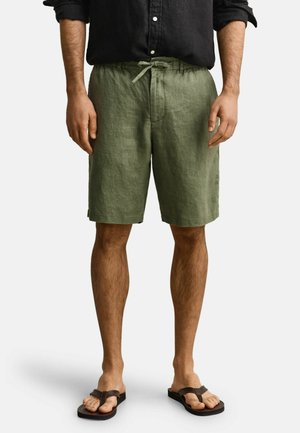 Man draagt olijfgroene korte broek met trekkoord, zwart overhemd met opgerolde mouwen en zwarte teenslippers, staand tegen een effen achtergrond.