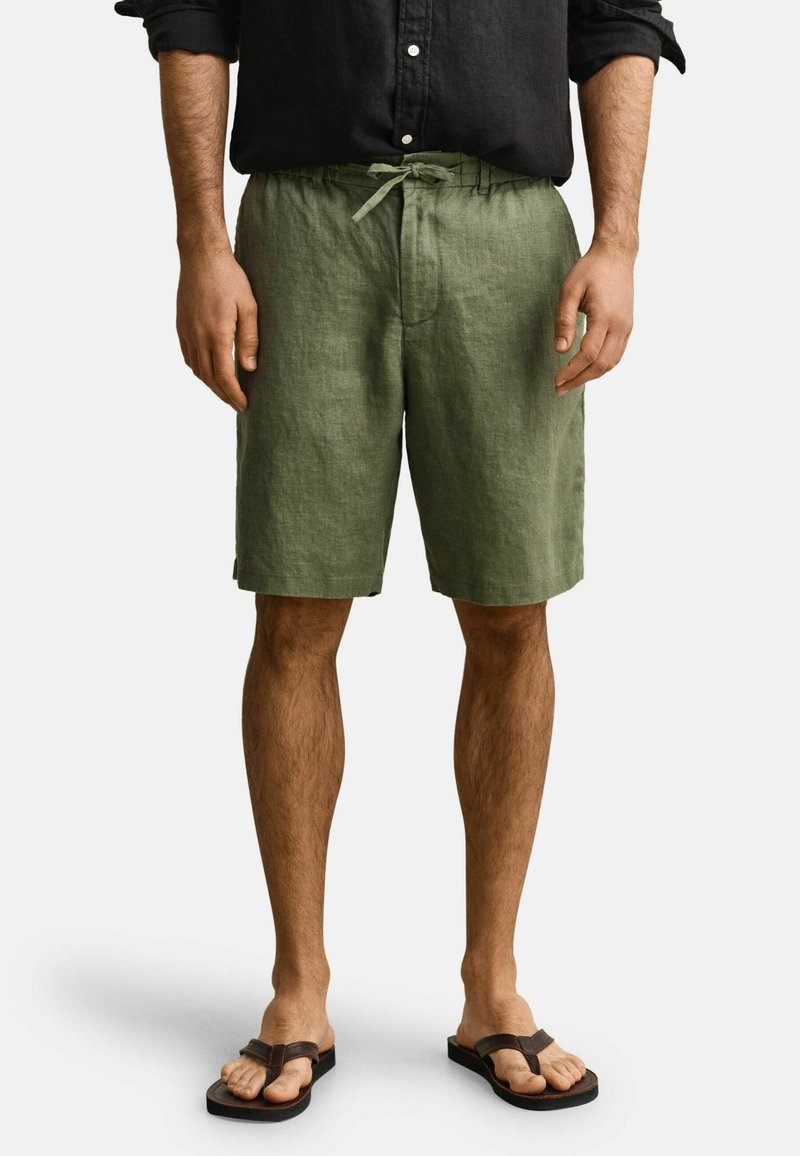 Homme portant un short à cordon olive, une chemise noire boutonnée aux manches retroussées et des tongs noires, debout devant un fond uni.
