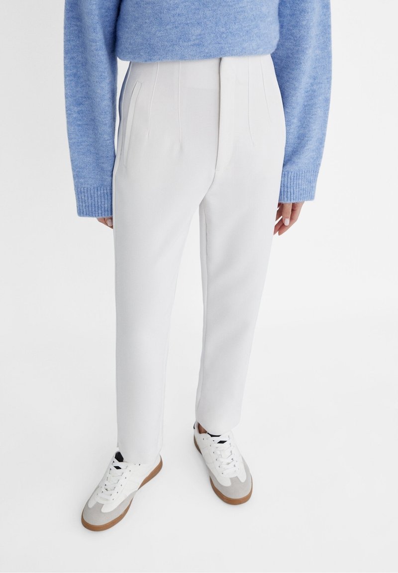 Personne portant un pull bleu clair, un pantalon blanc taille haute et des baskets blanches avec des semelles beige, debout sur fond blanc.