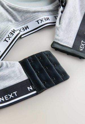 Tela gris con banda elástica negra y blanca que presenta el texto "NEXT". Detalle de cierre ajustable tipo gancho y ojal en negro visible. Textura suave.