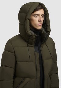 Olivenfarvet pufferjakke med hætte, der har et quiltet design, lynlås foran og justerbare snore ved halsen.
