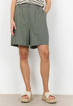 Shorts en lin verts avec une coupe décontractée, poches latérales et fermeture à bouton. Associés à des sandales espadrilles de couleur claire.