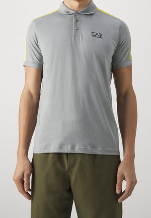 Homme portant un polo gris clair avec des rayures jaunes sur les épaules et le logo EA7 sur la poitrine gauche, associé à un pantalon vert olive.