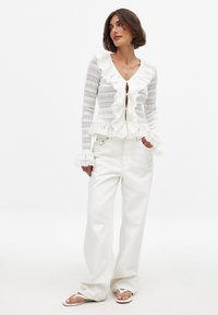 Gilet blanc à volants avec un col en V, fermeture à boutons et tricot texturé. Associé à un pantalon blanc ample et des tongs, design minimaliste.