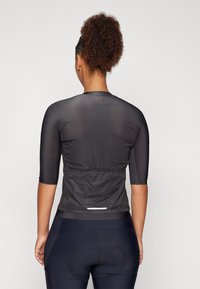 Schwarzes Sportoberteil mit kurzen Ärmeln, körperbetonter Passform, leichtem Stoff und gerafftem Taillenbereich. Getragen mit dunklen Leggings, die gepolsterte Akzente aufweisen.