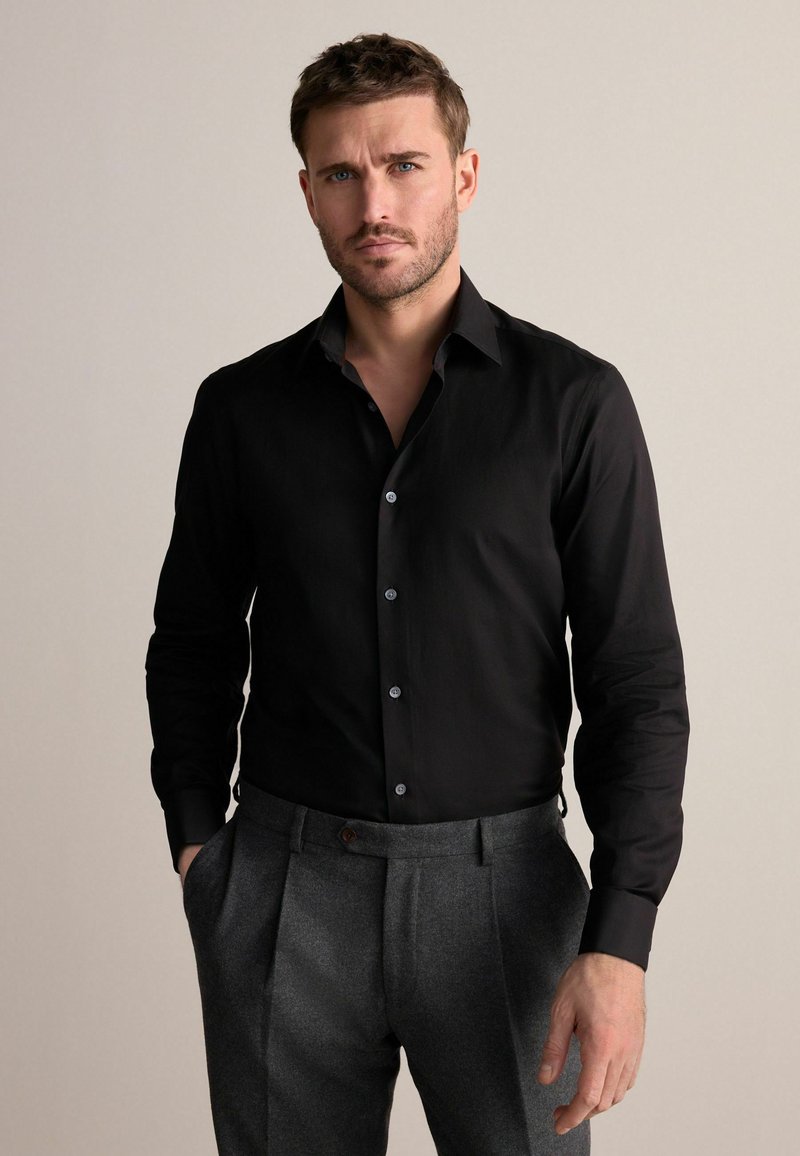 Schwarzes Langarmshirt mit Kragen, das über Knopfdetails und eine glatte Textur verfügt, kombiniert mit grauen gerade geschnittenen Hosen.