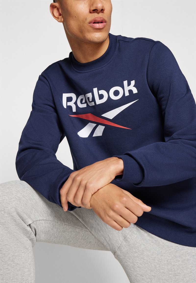 Granatowy sweatshirt z białym i czerwonym logo Reebok, z okrągłym dekoltem i ściągaczami przy rękawach, w połączeniu z jasnoszarymi spodniami dresowymi.