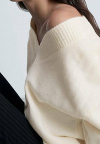 Pull crème à épaules dénudées avec un col côtelé, fabriqué en tissu doux, associé à un vêtement noir texturé.