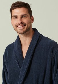 Tums zilā terijas halāts ar šahtu apkakli, teksturētu audumu un atvērtu priekšpusi, ko valkā smaidoša persona uz neitrāla fona.