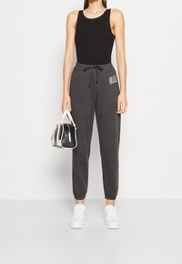 Frau trägt ein schwarzes Tanktop und dunkle graue GAP-Sweatpants, hält eine schwarz-weiße Handtasche in der Hand und steht mit weißen Turnschuhen vor einem einfarbigen Hintergrund.