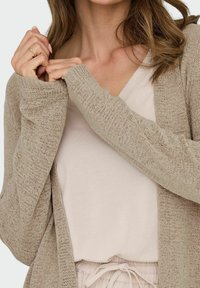 Cardigan tricoté taupe clair à manches longues, avec une coupe ample et un devant ouvert, superposé sur une chemise beige clair à col en V.