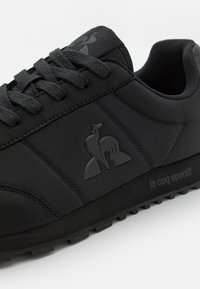 Scarpe da ginnastica nere con una tomaia sintetica liscia, accenti testurizzati e una suola in gomma con battistrada. Presentano una patta con logo sul lato e lacci.