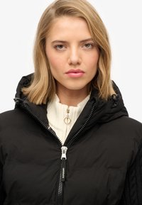 Zwarte pufferjack met een rits aan de voorkant, voorzien van een capuchon en kabelgebreide textuur op de mouwen. Draagt over een crèmekleurige geribbelde trui.