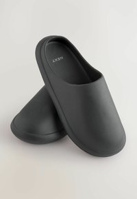Svarta slip-on skor i slät material, med en minimalistisk design, rundad form och texturerad innersula. Varumärket "NEXT" synligt.