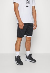 Shorts de sport noirs avec des bandes latérales, associés à un T-shirt blanc arborant un logo NBA noir, des chaussettes blanches et des chaussures de sport noires.