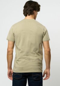 Mann med beige kortermet t-skjorte og mørkeblå jeans, vist fra bakgrunnen mot en ensfarget lys bakgrunn.