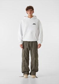 Biała bluza z kapturem i kieszenią z przodu, z czarnym logo, w połączeniu z oliwkowymi spodniami cargo z wieloma kieszeniami i luźnym krojem.