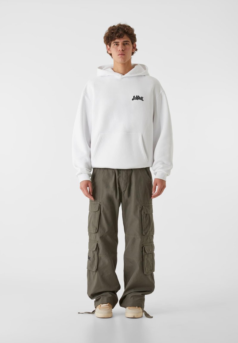 Biała bluza z kapturem i kieszenią z przodu, z czarnym logo, w połączeniu z oliwkowymi spodniami cargo z wieloma kieszeniami i luźnym krojem.