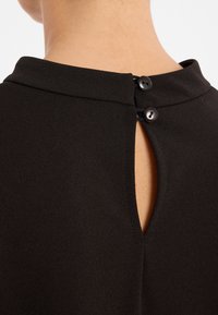 Schwarzer Stoff mit glatter Textur zeigt eine kleine Schlüssellochöffnung auf der Rückseite, die mit zwei schwarzen Knöpfen gesichert ist.