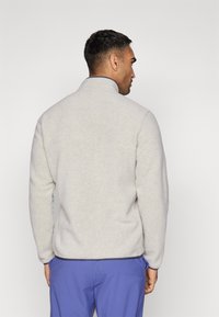 Jachetă de fleece gri deschis, cu un guler înalt și o textură fină. Dispune de un închidere cu fermoar și margini închise la culoare de-a lungul tivului și gulerului.