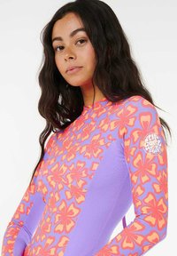 Camisa de manga larga con protección solar que presenta un vibrante patrón floral en rosa y naranja sobre un fondo morado, con un logo en la manga derecha.