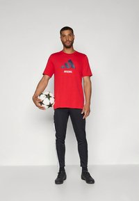 Rød atletisk t-shirt med "Arsenal"-logo, kombineret med sorte træningsbukser med hvide sidelister og sort fodtøj, holder en fodbold.