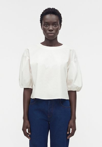 Femme aux cheveux courts et bouclés portant un chemisier blanc ample à manches courtes et un jean bleu foncé taille haute sur un fond uni.