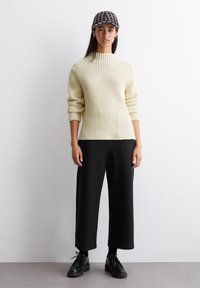 Cremfarbener Strickpullover mit hohem Kragen, kombiniert mit schwarzen weiten Hosen und schwarzen Schuhen. Das Accessoire besteht aus einer schwarz-weißen Mütze mit Muster.