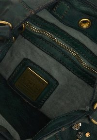 Interno di borsa in pelle verde con finiture dorate, fodera in tessuto, tasca con cerniera e un'etichetta che dettaglia informazioni sul marchio e sulla produzione.