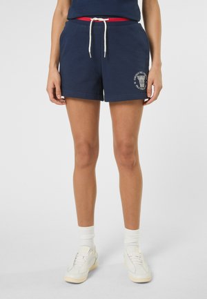 Osoba ubrana w granatowe spodenki z białym logo na lewej udzie, białe skarpetki i białe tenisówki, stojąca na jednolitym tle.