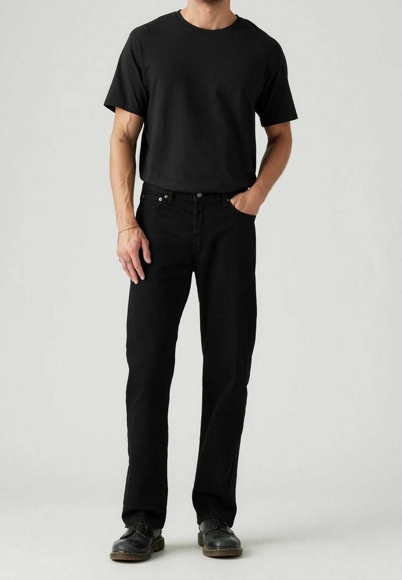 Homme portant un t-shirt noir uni, un jean noir et des chaussures noires, debout avec une main dans la poche devant un fond uni.
