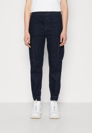 Persona con pantalones tipo jogger de mezclilla azul oscuro con bolsillos laterales y zapatillas blancas, de pie contra un fondo claro y liso.