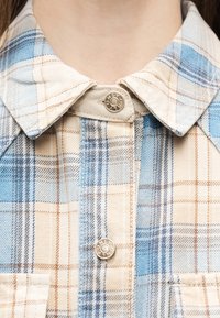 Κοντινό πλάνο του γιακά πουκαμίσου με καρό μοτίβο σε αποχρώσεις μπεζ, μπλε και καφέ, με ασημένια κουμπιά τύπου σναπ που φέρουν την επιγραφή "ONLY", στο λαιμό και το πάνω μέρος του στήθους ενός ατόμου.