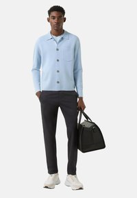 Lyseblå cardigan med knaplukning og brystlomme, stylet med mørke bukser og hvide sneakers, holder en sort taske.