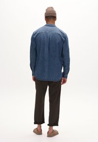 Jeanshemd in mittlerem Blau, lockere Passform mit langen Ärmeln und Rückenfalten. Getragen mit olivgrünen, hochgekrempelten Hosen und beigen Slippern.