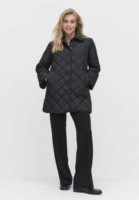 Svart vadderad jacka med knäppning framtill, spetsig krage och sidofickor; kombinerad med svarta vida byxor.