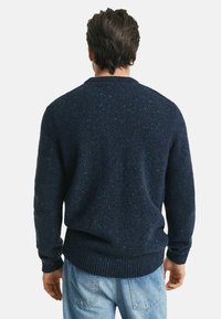 Uomo che indossa un maglione a maglia blu scuro con microdettagli e jeans blu chiaro, in piedi con le spalle rivolte verso uno sfondo bianco semplice.