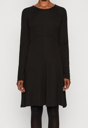 Femme vêtue d'une robe noire à manches longues, longueur genou, debout, bras détendus le long du corps, devant un fond uni.