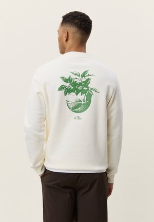 Homme de dos portant un sweat-shirt écru avec une plante verte poussant d’un graphisme de basket-ball fissuré dans le dos, associé à un pantalon sombre.