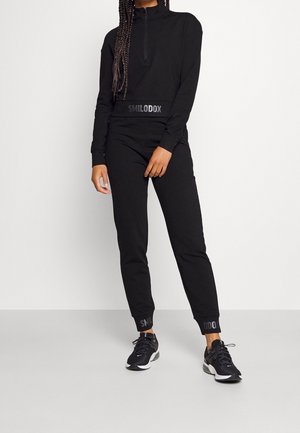 Frau trägt schwarzen Sweatshirt mit Reißverschluss und passende Jogginghose mit "SMILODOX"-Markenaufdruck, kombiniert mit schwarzen Sportschuhen, steht vor weißer Wand.